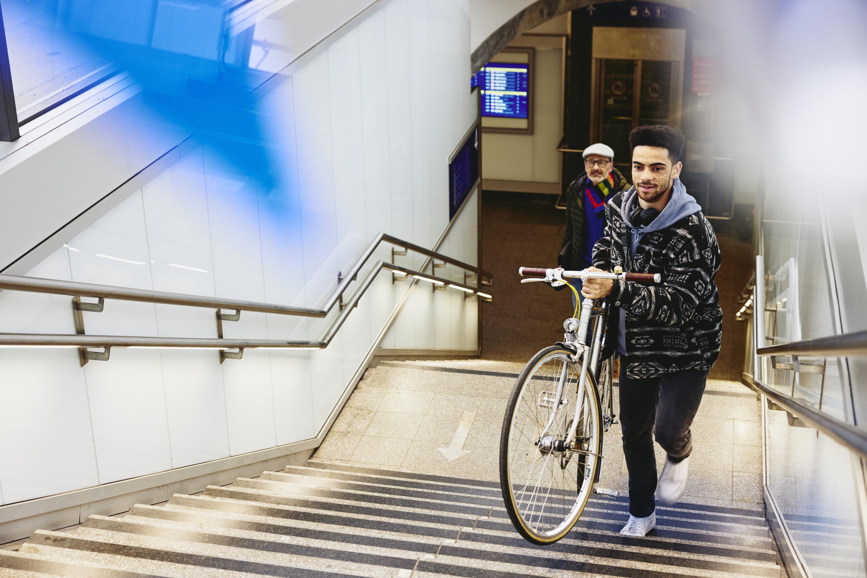 Man carrying a bike up the stairs in a train station. Other man walking up the stairs in the background.