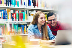  A woman with long, blonde hair and a denim shirt smiles while looking at a laptop. A man with short, dark hair, glasses, and a red hoodie is looking at the laptop over their shoulder, also smiling. They appear to be in a library, with bookshelves filled with books in the background.