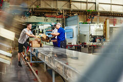 Three workers are in a warehouse, moving boxes on a conveyor belt. One worker in a grey shirt hands a box to another, while a third worker watches. The background shows factory machinery.