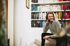 Smiling man sitting on stairs, having a phonecall. bookshelf in background