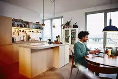 Smiling male sitting at his dining table working on his laptop. Kitchen in the background.
