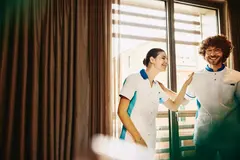 Two colleagues smiling, working housekeeping in a hotel room.
