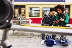male and female looking at phone while sitting on a bench on a train platform.
