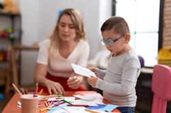 Female teaching assistant helping a SEN child with a crafts project, sat at a table using pens and paper.