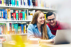  A woman with long, blonde hair and a denim shirt smiles while looking at a laptop. A man with short, dark hair, glasses, and a red hoodie is looking at the laptop over their shoulder, also smiling. They appear to be in a library, with bookshelves filled with books in the background.