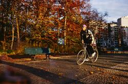 A person wearing a plaid jacket and a purple hat rides a bicycle quickly through a park setting in autumn. The trees are filled with bright orange and yellow foliage.