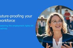 An eye-level shot captures a smiling woman with glasses and blonde hair, dressed in a black blazer and a white top, against an outdoor backdrop. She is positioned slightly to the right, engaged in conversation, with her hands raised as if explaining something. The background, softly blurred, features a person in a red shirt and white pants, along with what appears to be part of a building. The dominant colors in the image are blue, black, and white. The overall impression is one of professionalism.