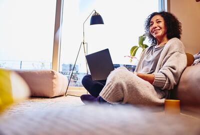 Smiling woman leaning against a couch, sitting on the floor, with laptop on her lap.