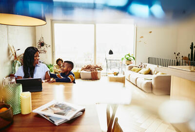 Woman working on tablet at the kitchen table. Kid sitting next to her. Male in the background.