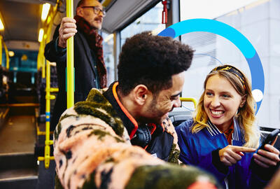 Man and woman looking at a phone and having a conversation on a bus. Other people in the background.