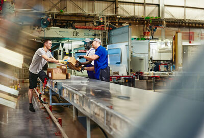 Three workers are in a warehouse, moving boxes on a conveyor belt. One worker in a grey shirt hands a box to another, while a third worker watches. The background shows factory machinery.