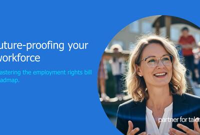 An eye-level shot captures a smiling woman with glasses and blonde hair, dressed in a black blazer and a white top, against an outdoor backdrop. She is positioned slightly to the right, engaged in conversation, with her hands raised as if explaining something. The background, softly blurred, features a person in a red shirt and white pants, along with what appears to be part of a building. The dominant colors in the image are blue, black, and white. The overall impression is one of professionalism.