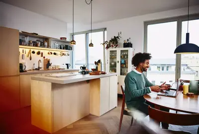 Smiling male sitting at his dining table working on his laptop. Kitchen in the background.
