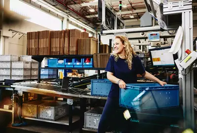 Smiling female standing next to conveyer belt holding a crate
