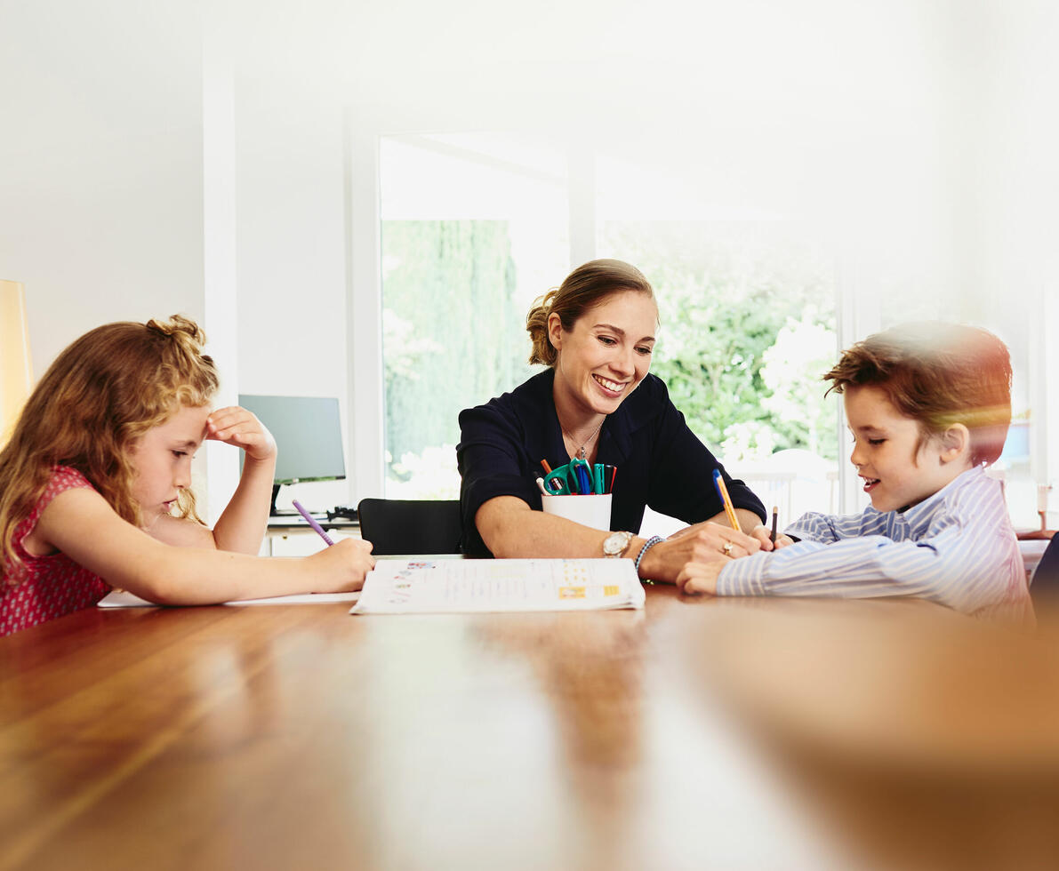 Smiling woman helping two kids at a dining table.