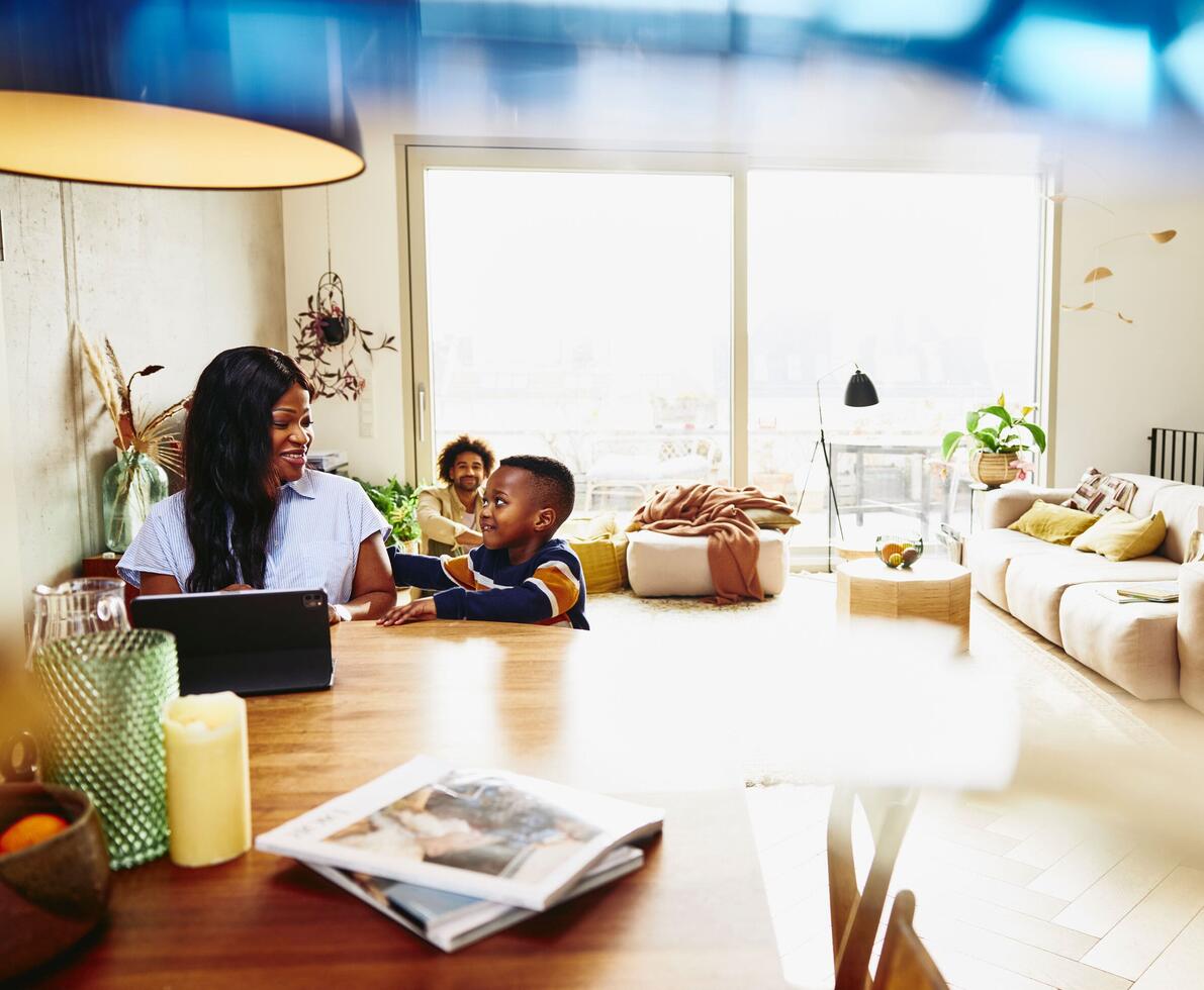 Woman working on tablet at the kitchen table. Kid sitting next to her. Male in the background.