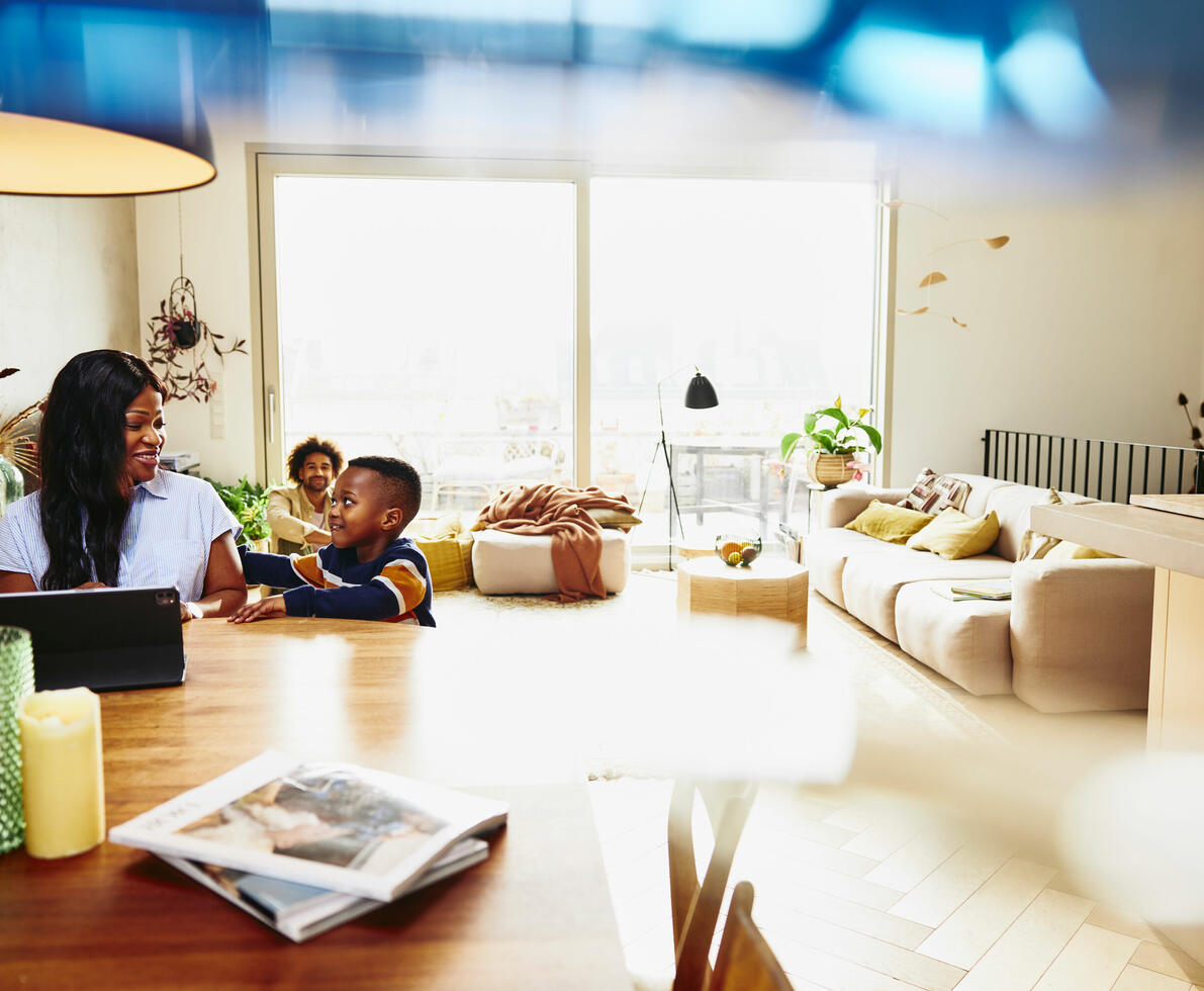 Woman working on tablet at a kitchen table. Kid sitting next to her. Man in the background.