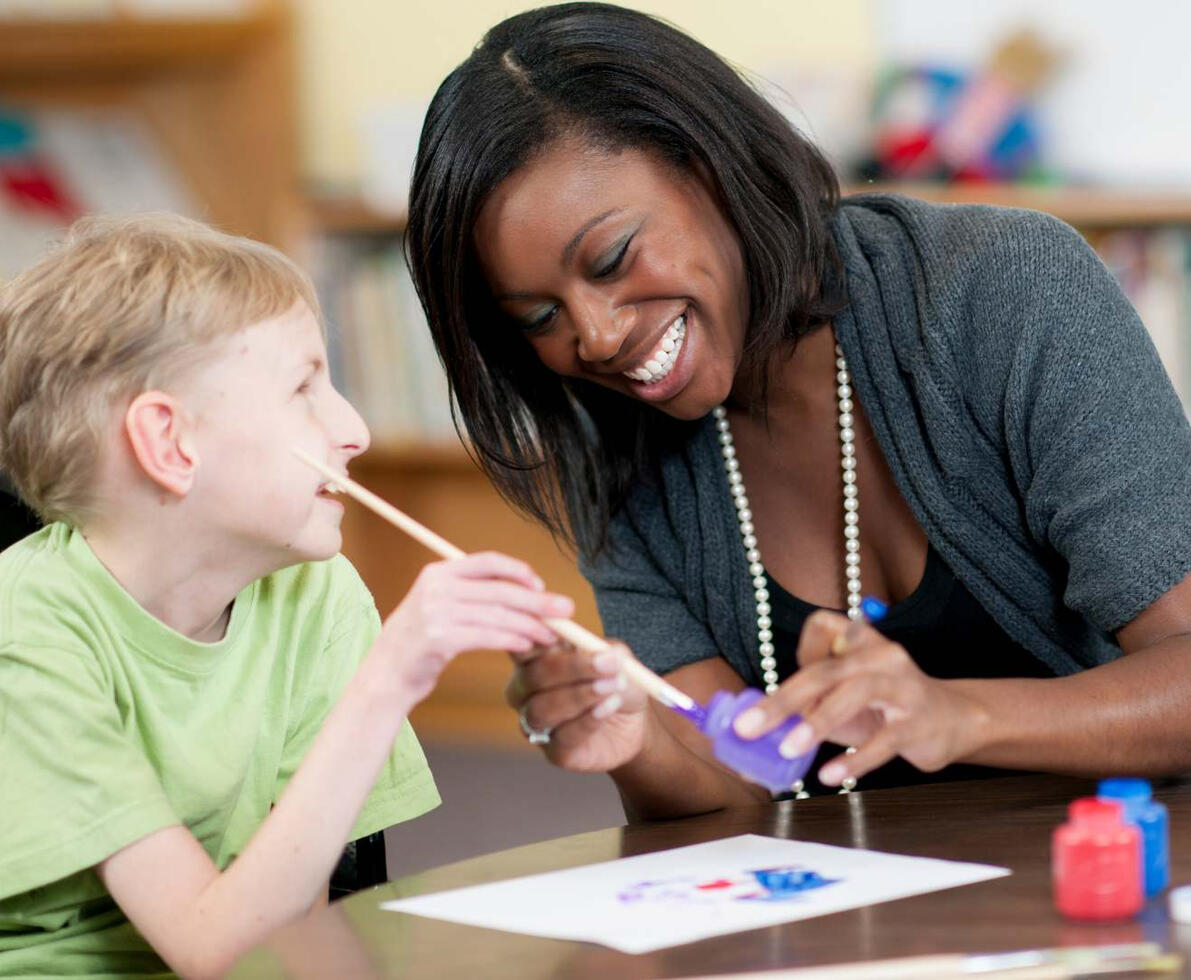Female teaching assistant helping a SEN child with a crafts project using paints.