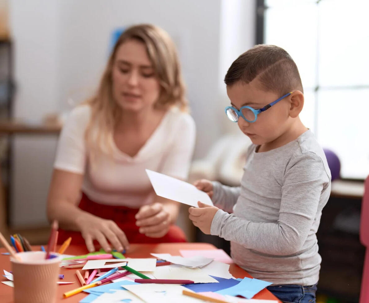Female teaching assistant helping a SEN child with a crafts project, sat at a table using pens and paper.