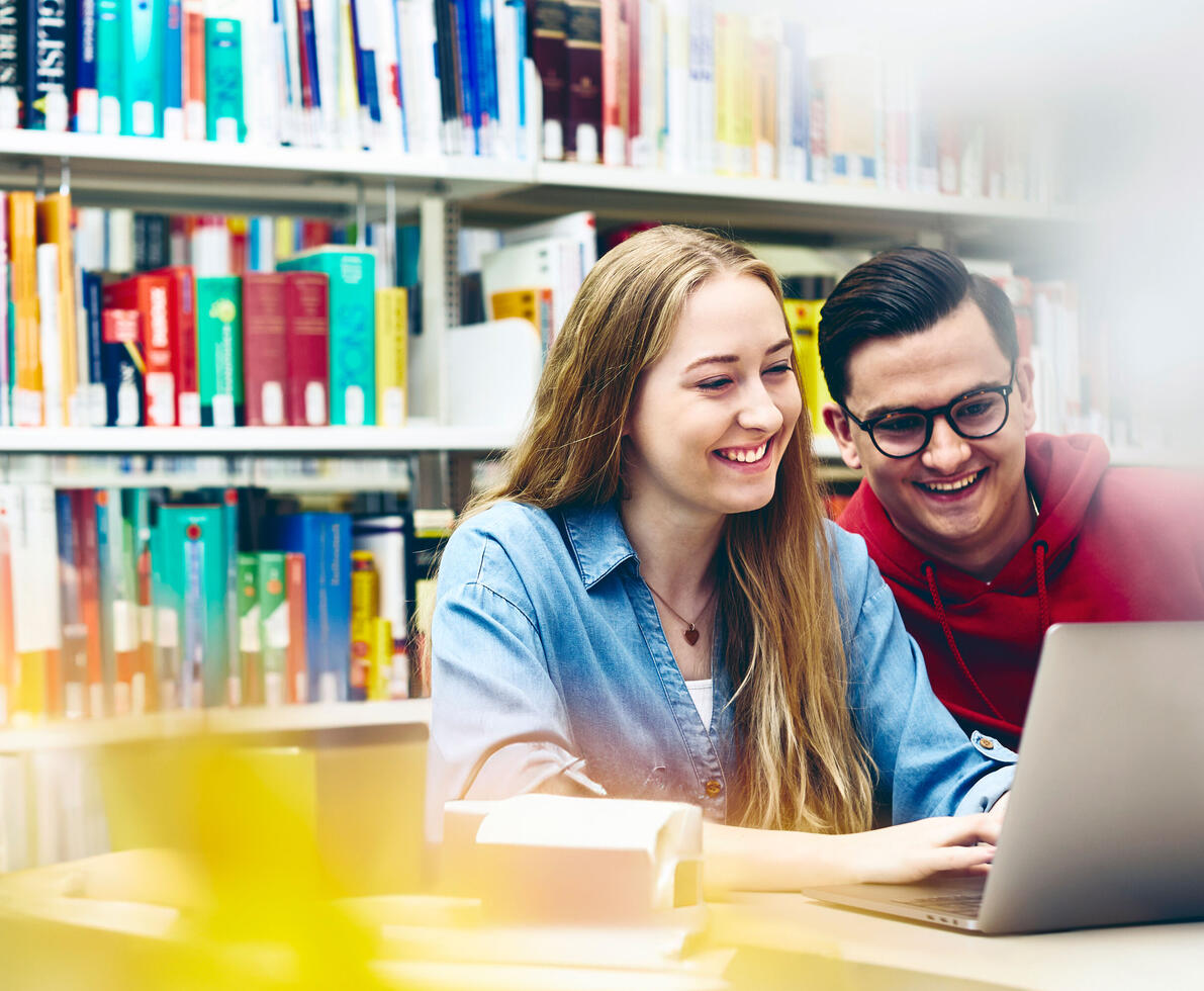  A woman with long, blonde hair and a denim shirt smiles while looking at a laptop. A man with short, dark hair, glasses, and a red hoodie is looking at the laptop over their shoulder, also smiling. They appear to be in a library, with bookshelves filled with books in the background.