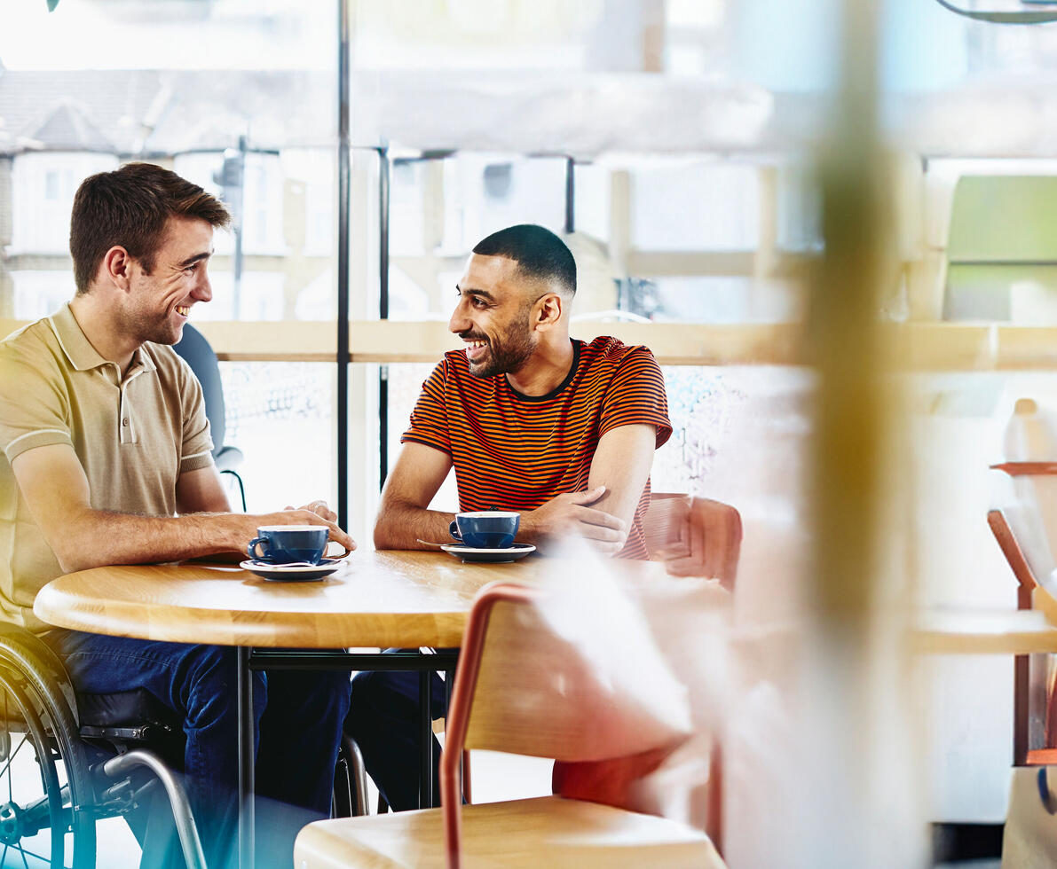 Two men are seated at a round wooden table in a cafe, facing each other and smiling, with drinks in front of them. The man on the left is in a wheelchair and is wearing a light-coloured polo shirt. The man on the right is wearing a striped t-shirt.
