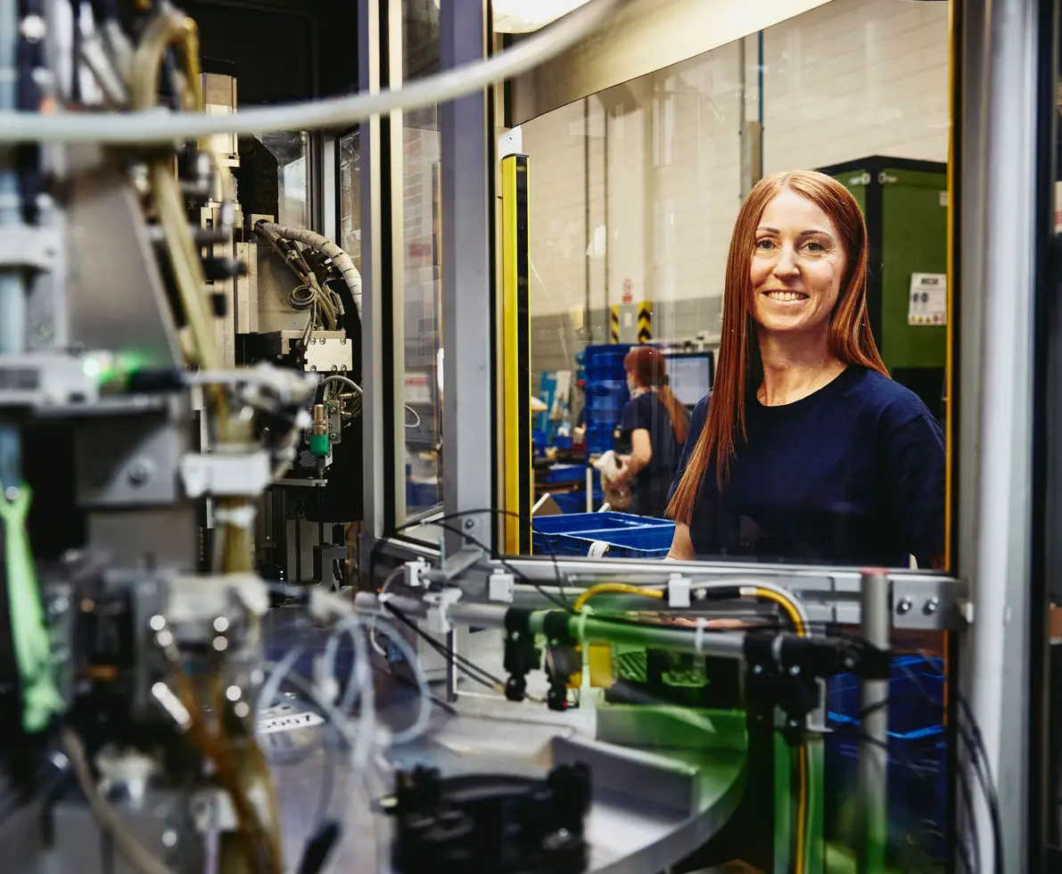 A red-haired woman in a dark blue shirt smiles at the camera in a factory setting. In front of her, the intricate parts of a machine are visible. In the background, another person works with blue plastic containers.