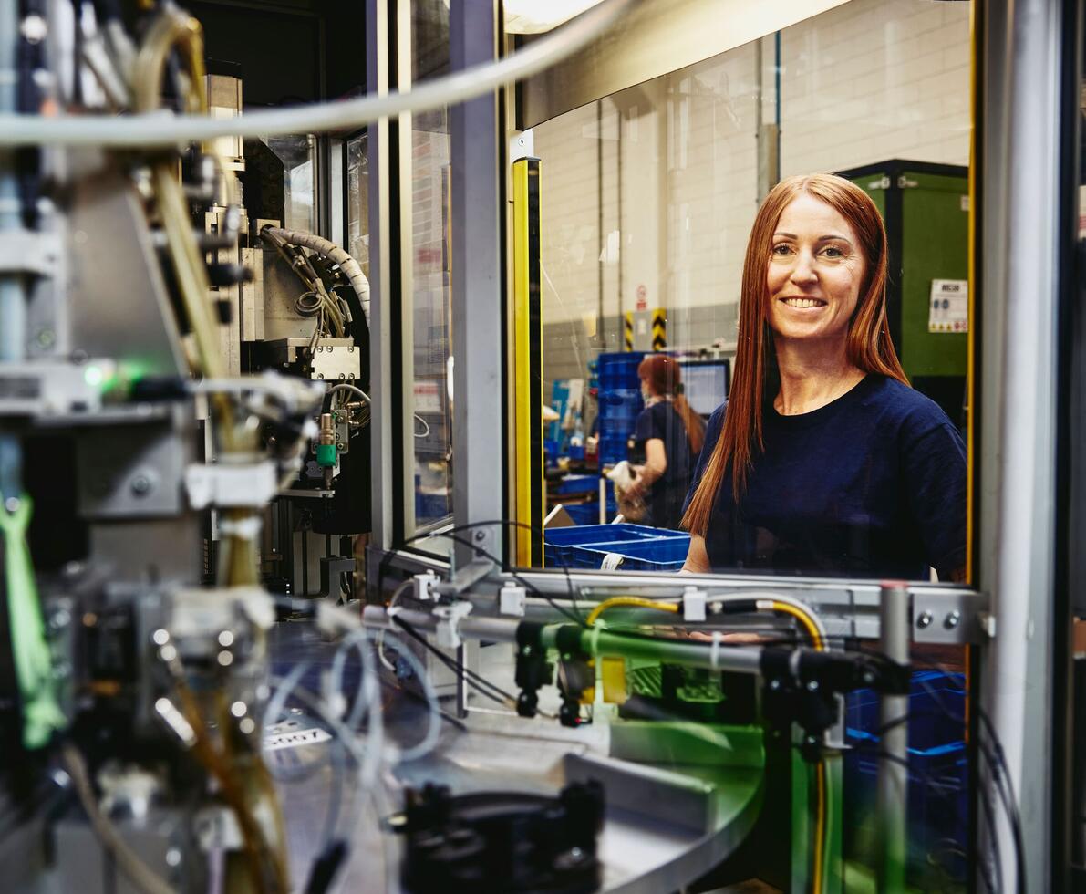 A red-haired woman in a dark blue shirt smiles at the camera in a factory setting. In front of her, the intricate parts of a machine are visible. In the background, another person works with blue plastic containers.