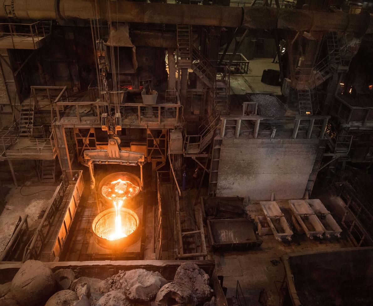  A high-angle shot inside a dark, industrial factory. A large, glowing crucible pours molten metal into a second, large container on the left, casting a bright orange light on the surrounding equipment and metal structures. In the foreground, there are piles of rubble and scrap metal.
