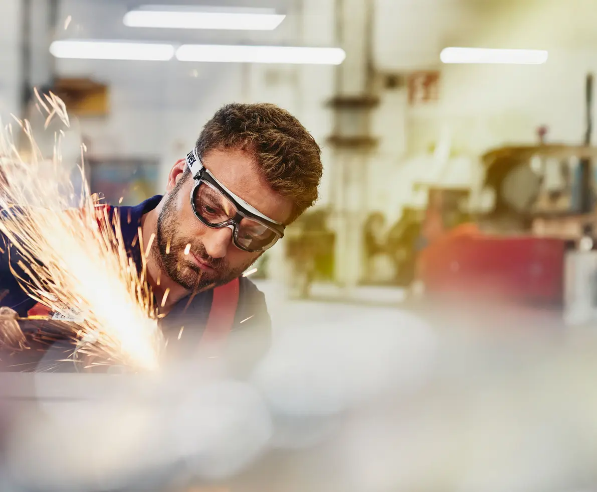 A man wearing safety goggles operates a grinding tool, creating a shower of bright sparks. He is working inside a factory. The background is blurred, drawing attention to the man and the sparks.