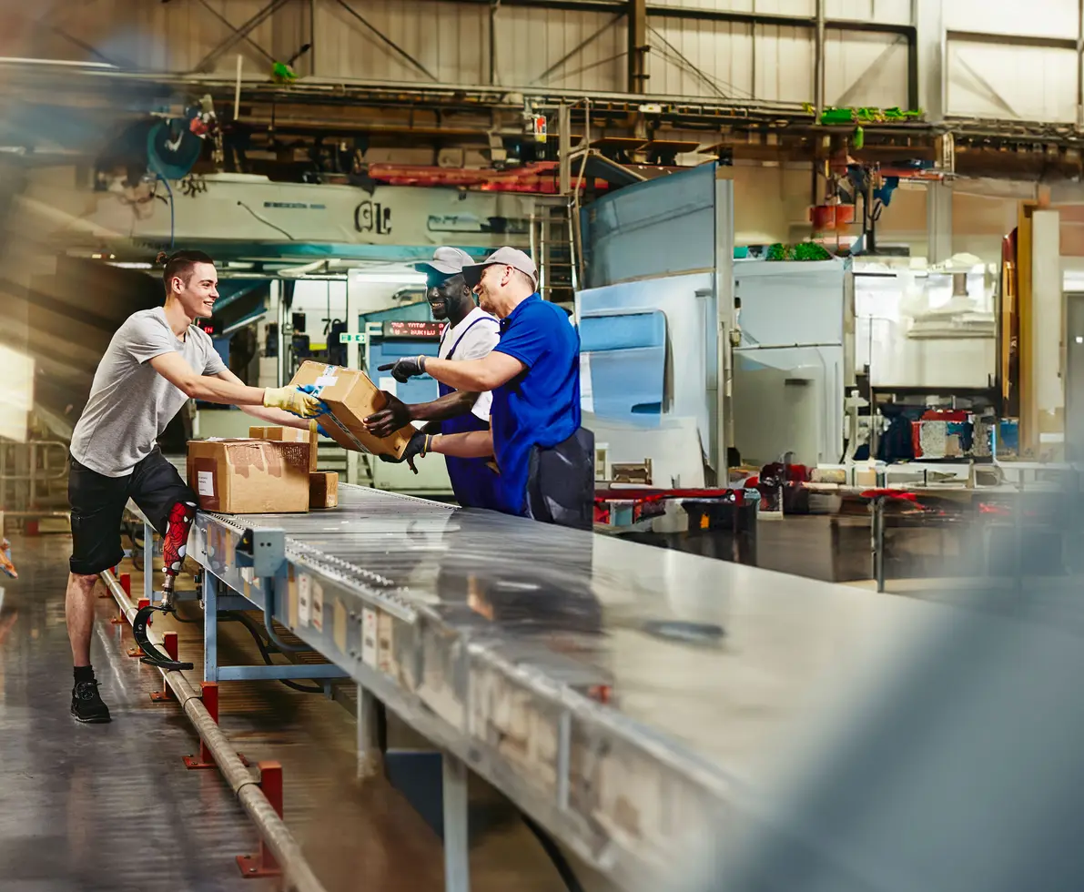 Three workers are in a warehouse, moving boxes on a conveyor belt. One worker in a grey shirt hands a box to another, while a third worker watches. The background shows factory machinery.
