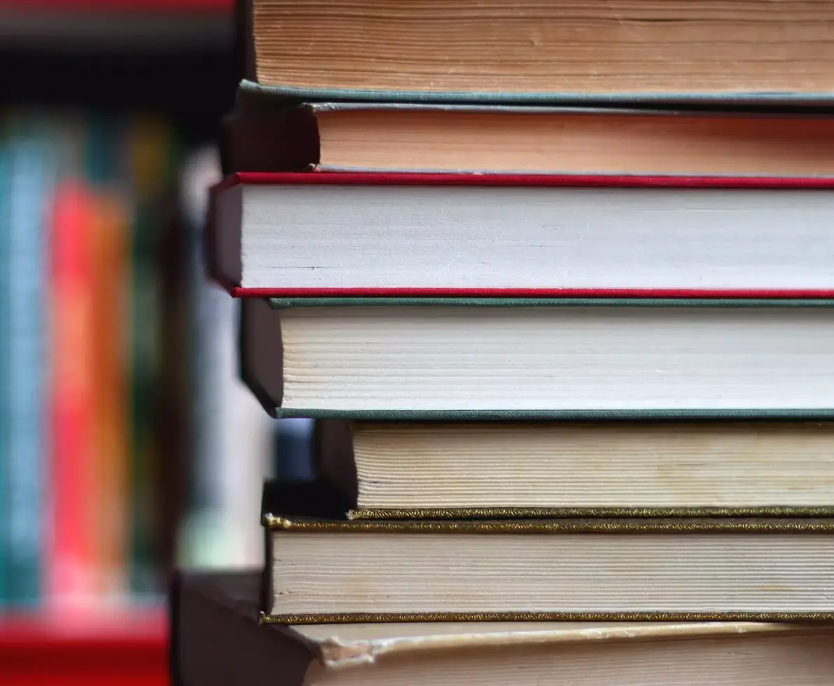 Stack of colorful hardcover books and reading glasses in front of a bookshelf. Selective focus.