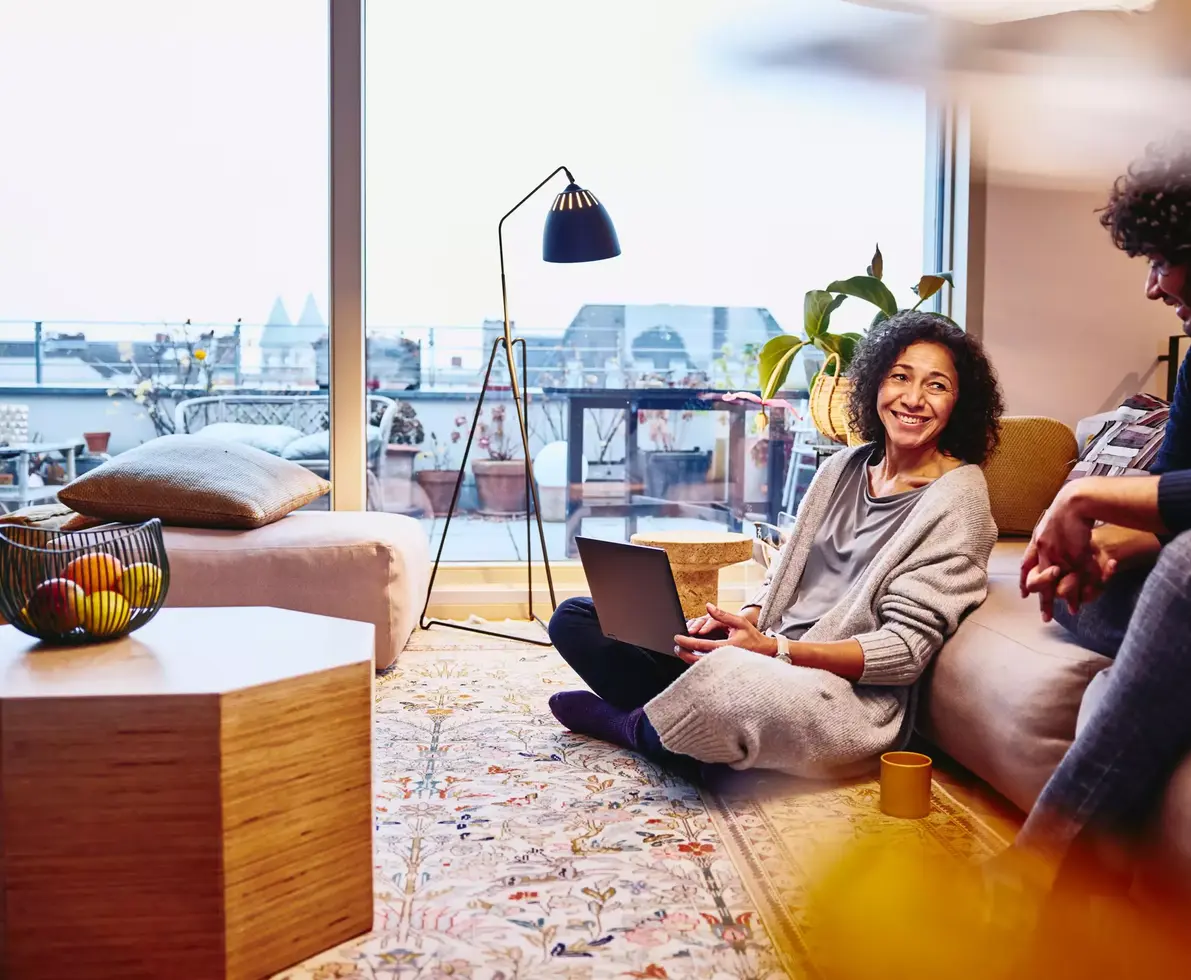 Smiling female leaning against a couch, sitting on the floor, with laptop on her lap. male sitting on couch.
