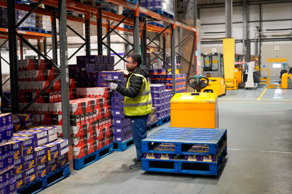  A male warehouse worker, wearing a high-visibility vest, is standing in an aisle of a large warehouse. He is looking at and scanning items on a pallet stacked with boxes of products. Tall shelves filled with more boxed goods line the aisle. In the background, other yellow warehouse machinery is visible.