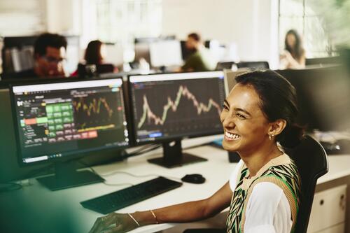 Woman smiling while sitting behind her desk, screens displaying financial information.