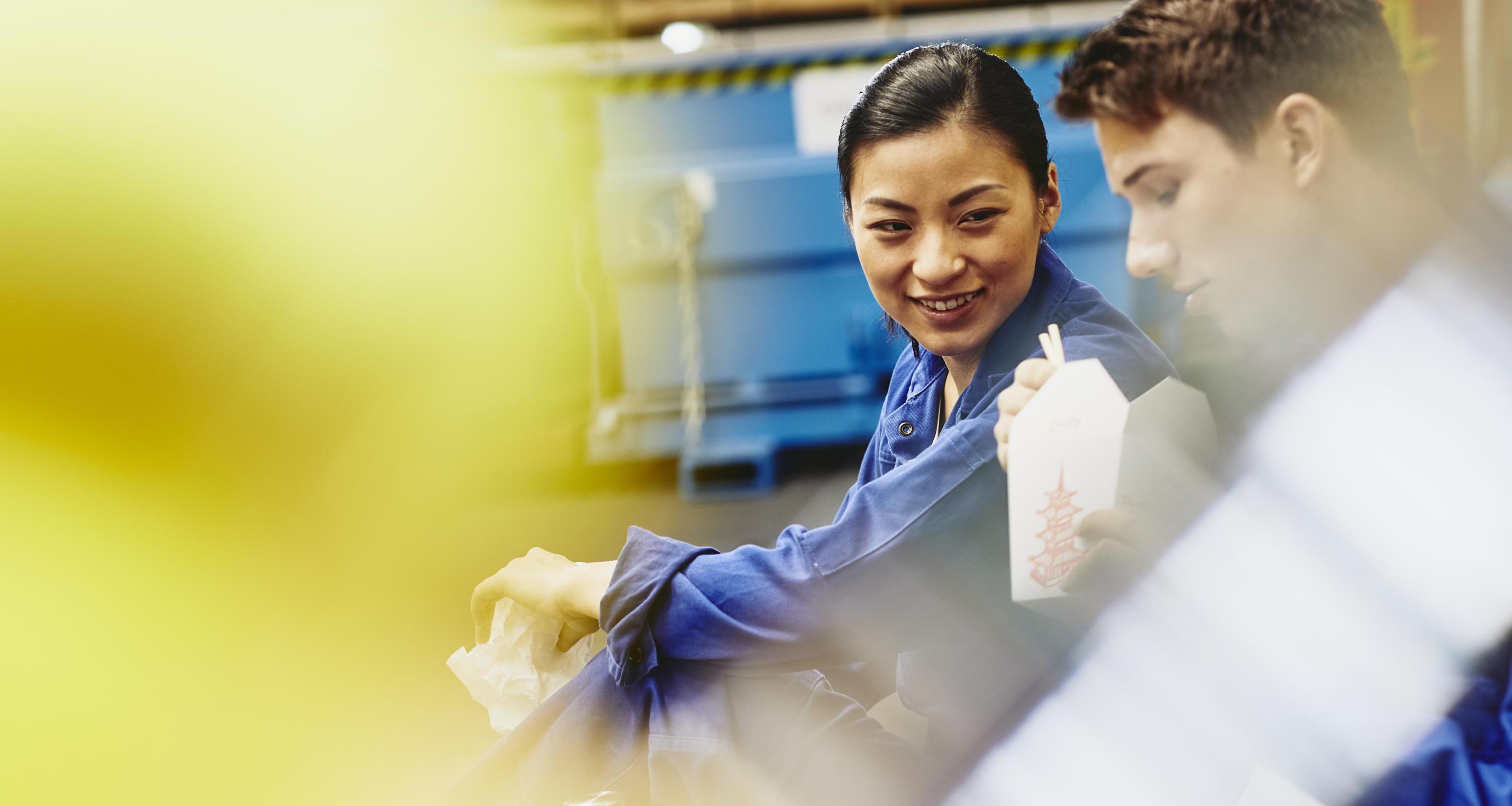 Male and female operator eating together. Food with chopsticks. Tech environment. Factory. Caucasian man and Asian woman. Primary color yellow. Secondary colors blue and white.