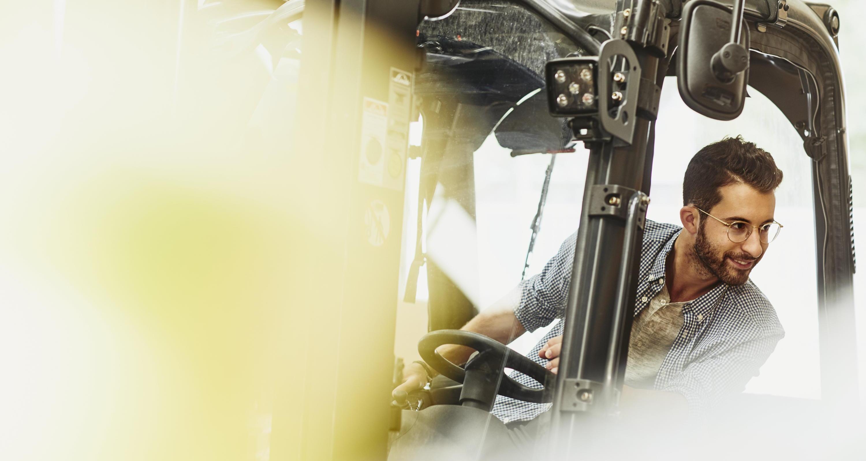 Male caucasian worker operating a forklift in a logistics environment. Looking to his left. Blue-collar. Smiling.  Wearing glasses. Groomed beard and moustache. Checkered shirt. Primary color cream.