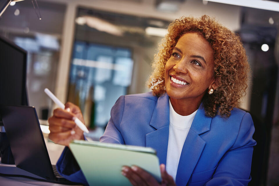Woman in a blue blazer, holding a touch pen and a tablet, sitting at a desk.