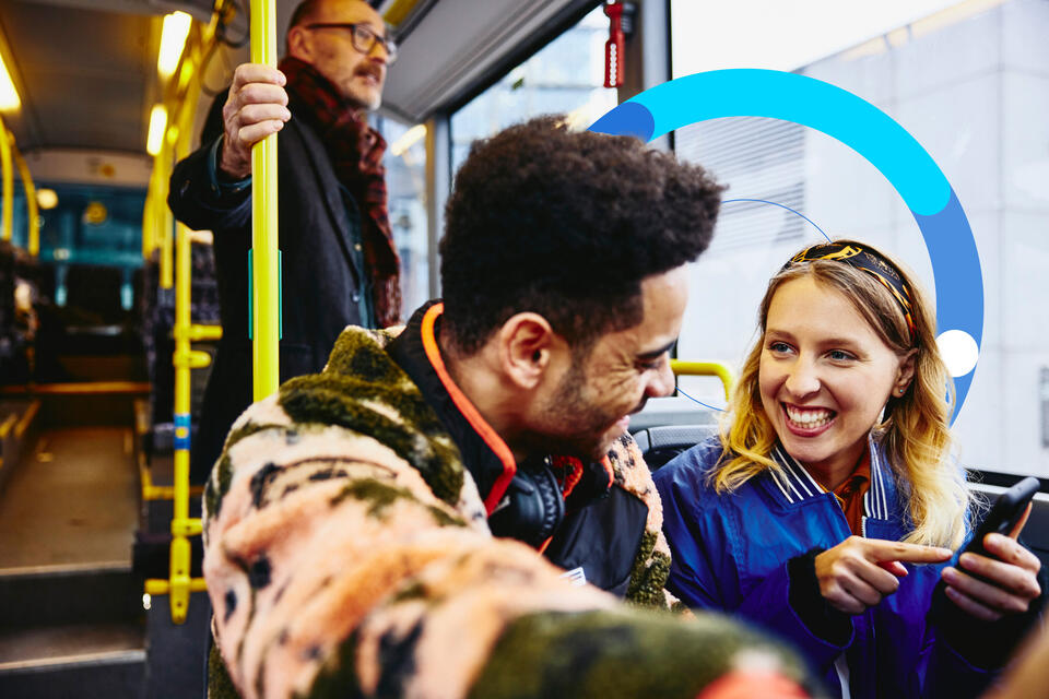 Man and woman looking at a phone and having a conversation on a bus. Other people in the background.