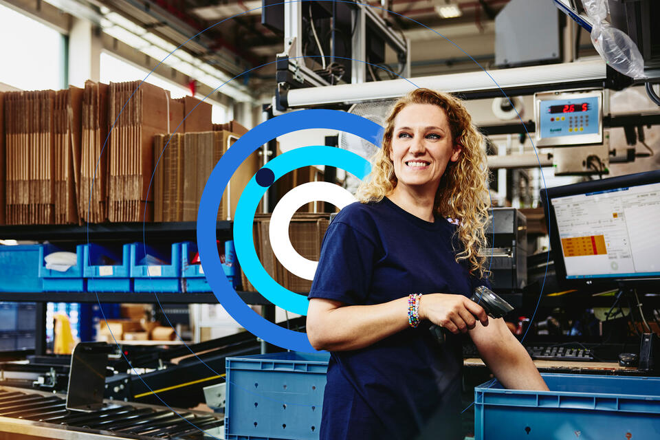 Woman smiling, standing next to a conveyer belt holding a scanner.