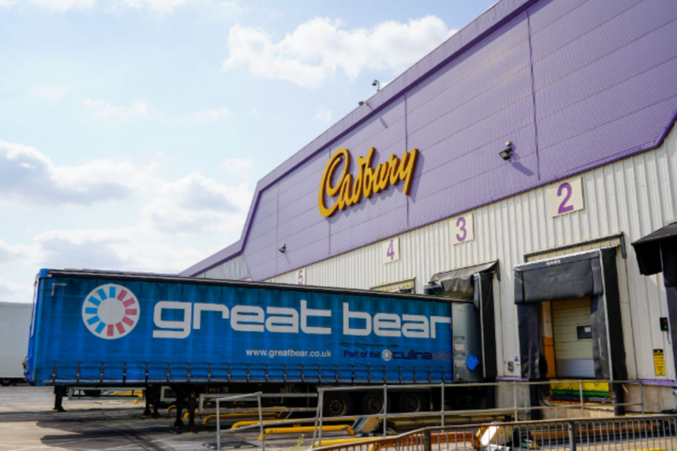A wide shot of the exterior of a large Cadbury warehouse. The building features a distinctive purple upper section with the yellow "Cadbury" logo, and a white lower section with numbered loading bay doors. A bright blue "great bear" logistics trailer is backed into one of the loading bays under a clear blue sky with some white clouds.