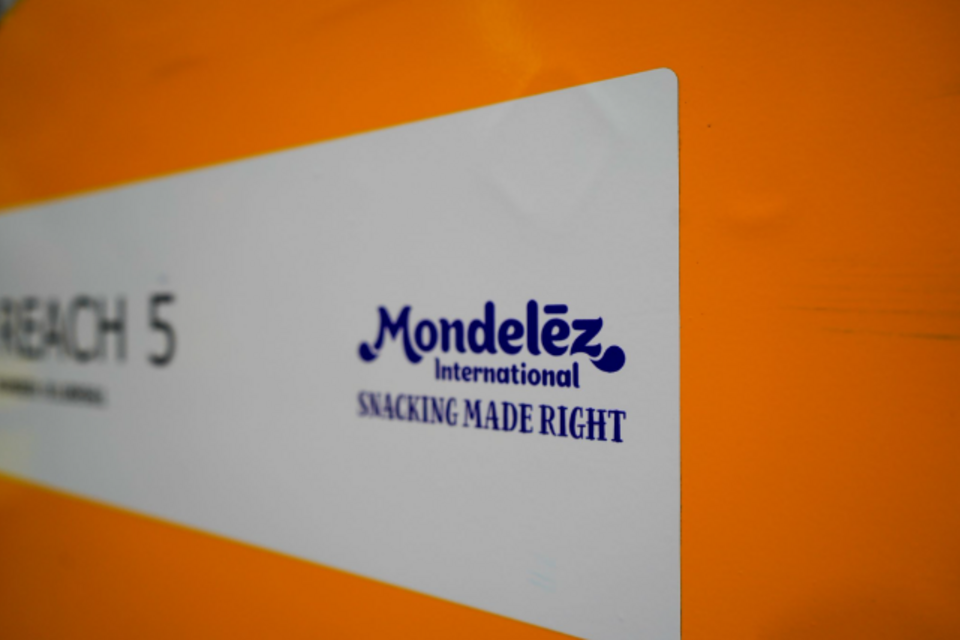  A close-up shot of a white sign with text, set against an orange background. The prominent text on the sign is the blue "Mondelez International" logo, with the slogan "SNACKING MADE RIGHT" underneath.