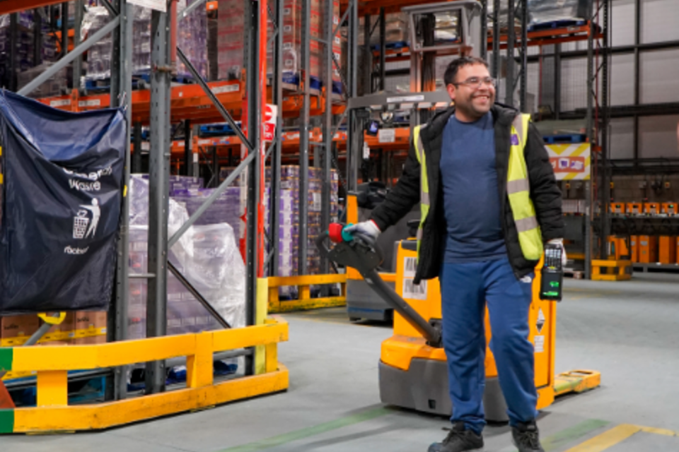 A man wearing a high-visibility vest and blue trousers is operating a pallet jack in a large warehouse. He is smiling and looking to his right. The warehouse has tall shelves stacked with various goods. 