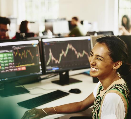 Woman smiling while sitting behind her desk, screens displaying financial information.